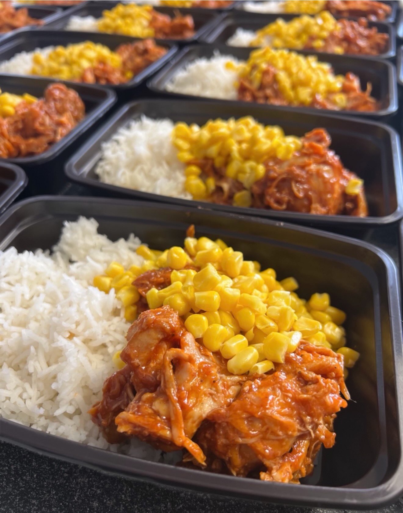 Rows of freshly prepped BBQ chicken bowls with basmati rice and corn in black meal-prep trays